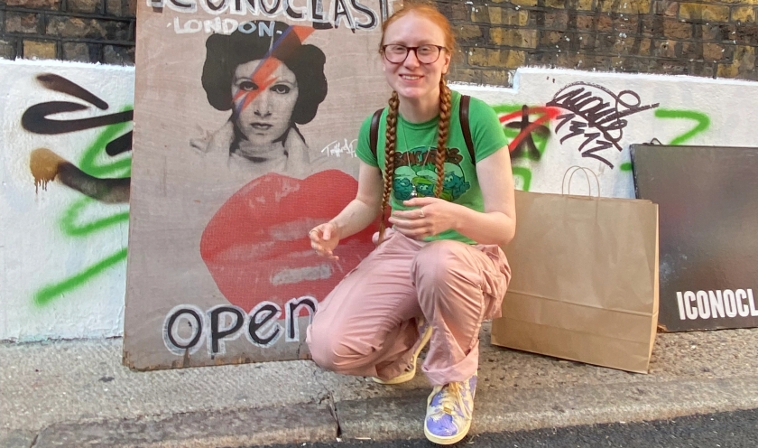 Millie, 19, is almost kneeling next to a sign against a brick wall. The signs has a picture of Princess Leia with Bowie's red and blue lightning bolt across her eye. Millie is wearing a green top, pink trousers, glasses, and her ginger hair is in plaits